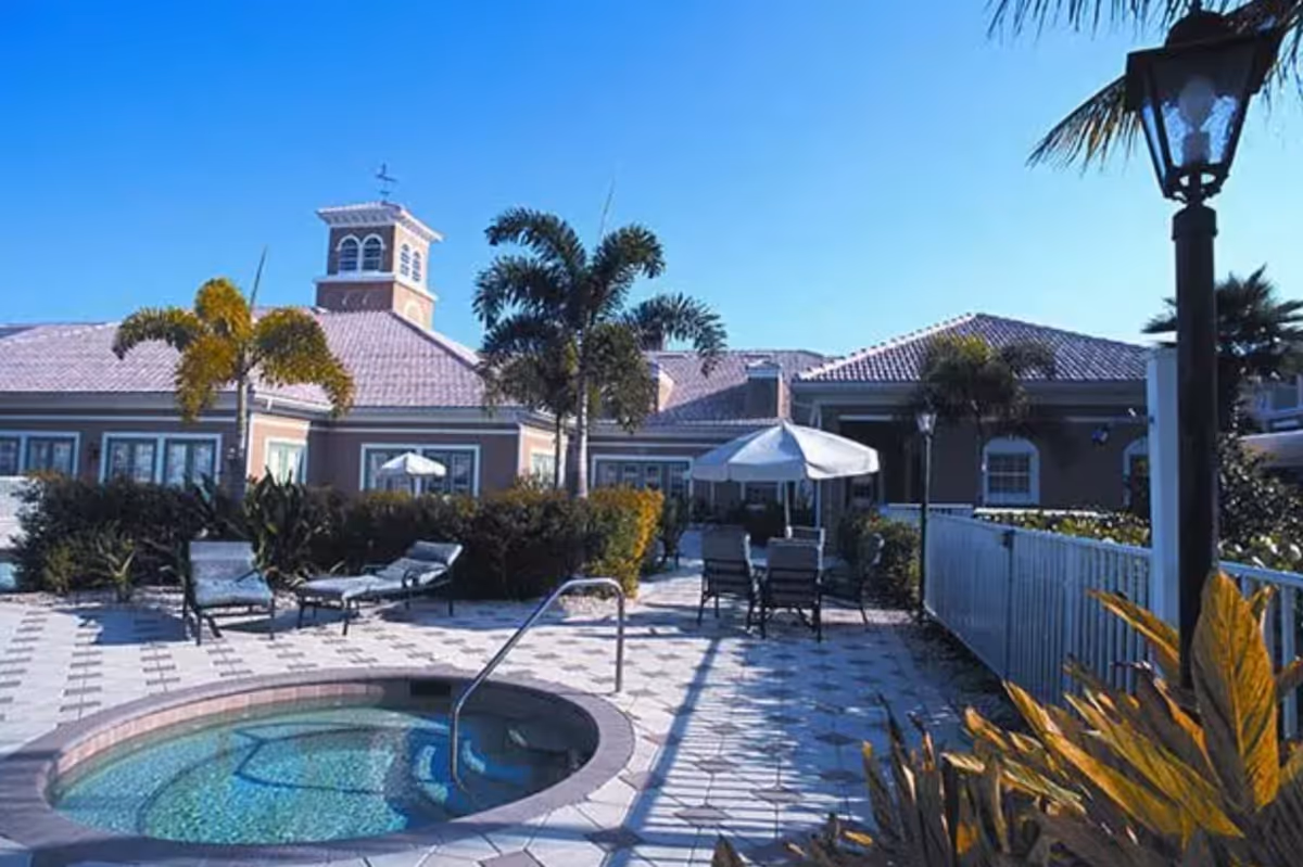 Outdoor patio area at Aston Gardens At Pelican Marsh featuring a small round pool or hot tub, lounge chairs, tables with umbrellas, palm trees, and a building with a cupola in the background under a clear blue sky.