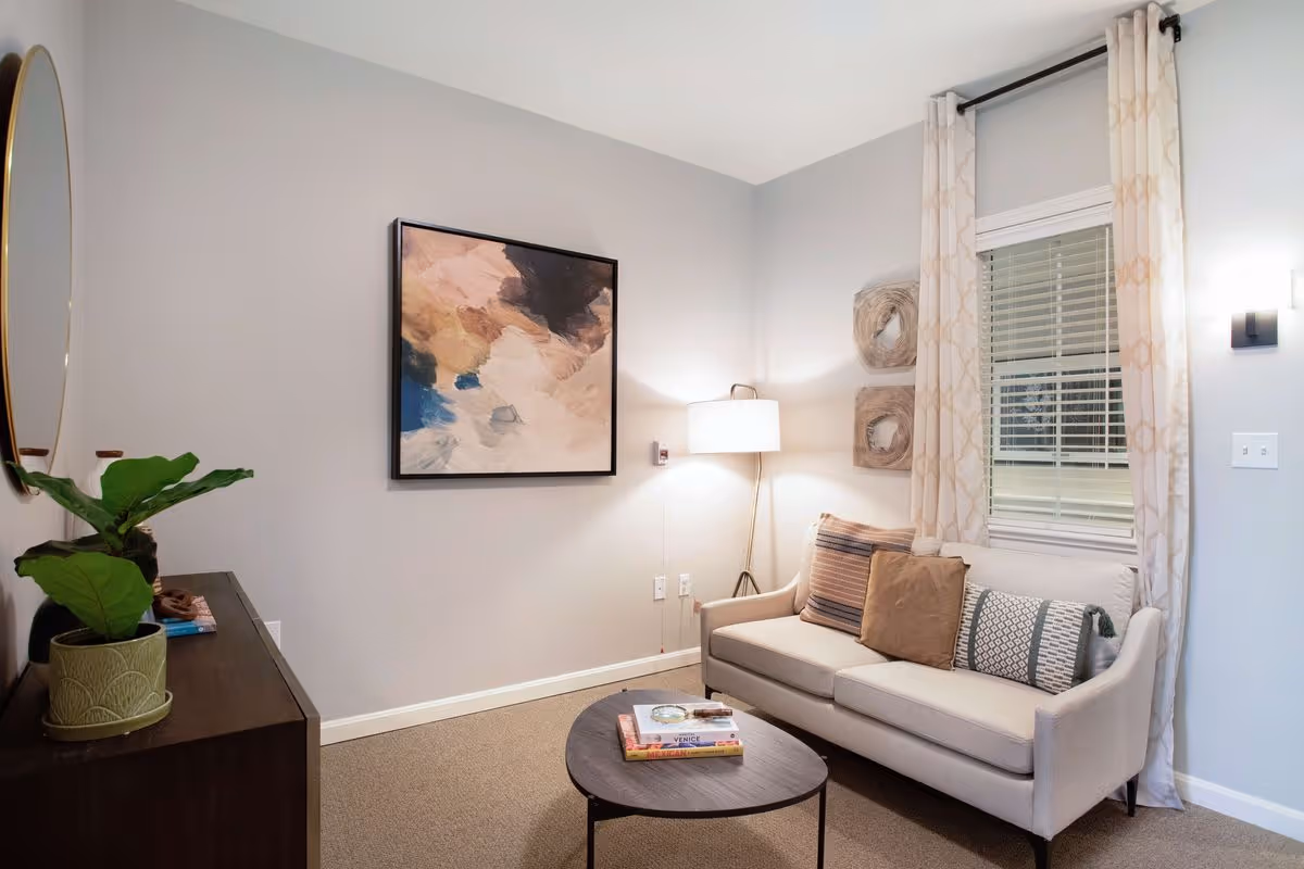 A cozy living room with a beige sofa adorned with three decorative pillows, a round wooden coffee table with books on it, a floor lamp in the corner, abstract artwork on the wall, a window with patterned curtains, and a dark wooden cabinet with a potted plant and a round mirror above it.