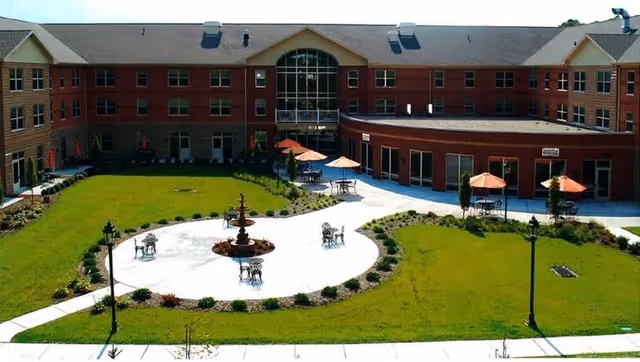Outdoor courtyard area of Paramount Senior Living Bethel Park featuring a circular paved patio with a central fountain, surrounded by tables with umbrellas and chairs, bordered by green lawns and landscaping, with a three-story brick building in the background.