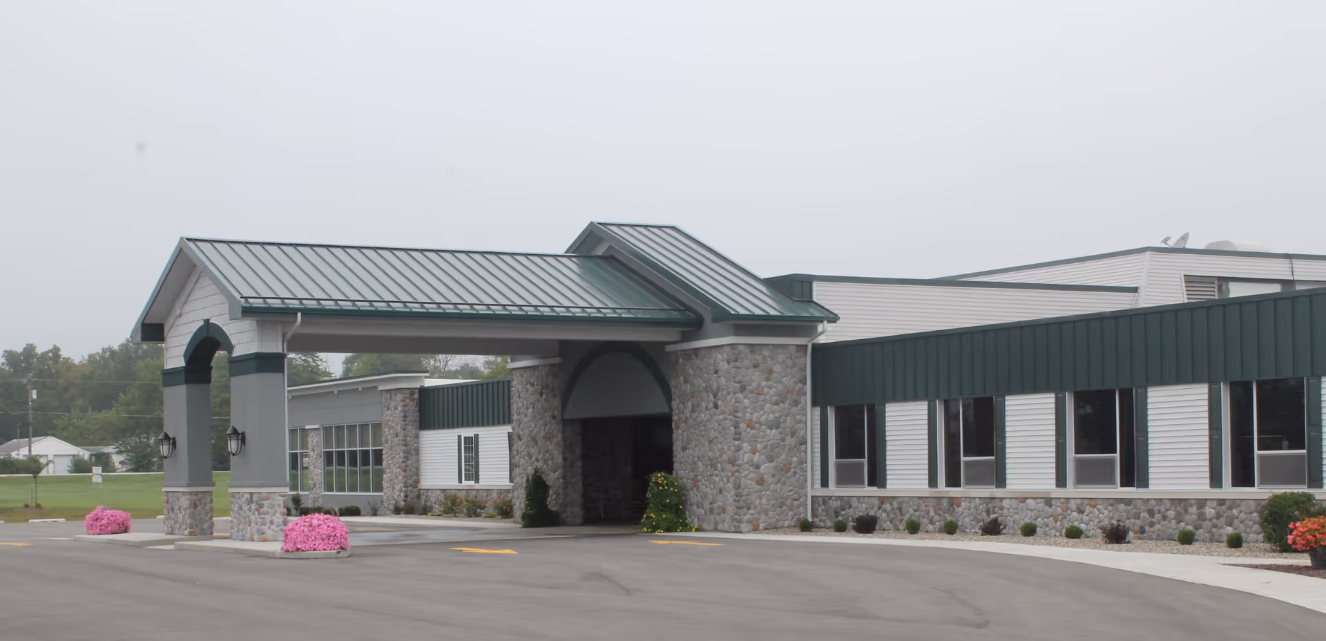 Exterior view of Miller's Merry Manor building featuring a covered entrance with stone pillars and a green metal roof, surrounded by a paved driveway and landscaped with flower beds.