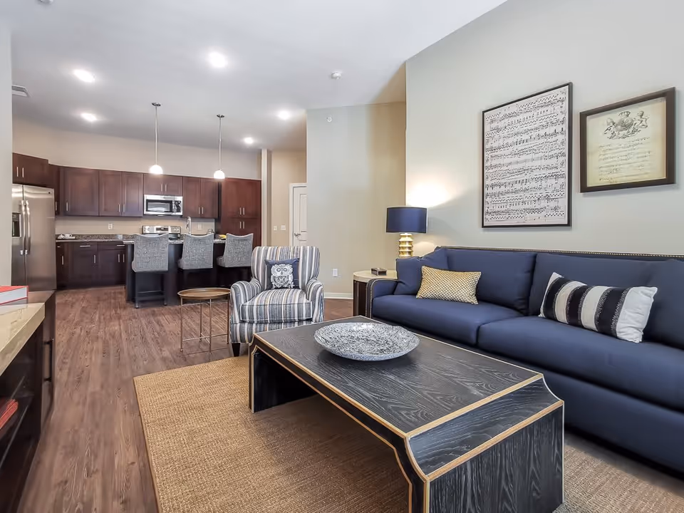 A modern living room and kitchen area in a senior living facility. The living room features a blue sofa with three decorative pillows, a striped armchair with an owl cushion, a black wooden coffee table with a decorative bowl, and a floor lamp with a blue shade. The kitchen area has dark wood cabinets, stainless steel appliances including a refrigerator and microwave, a granite countertop, and three gray upholstered bar stools at the kitchen island. The walls are light-colored with framed musical notes artwork hanging above the sofa. The floor is wood with a woven area rug in the living room.