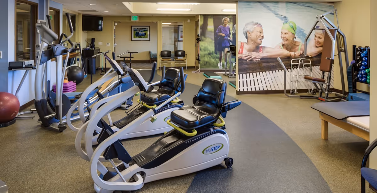 Interior fitness room with recumbent exercise machines, weight and therapy equipment, and a mural of smiling seniors on the wall.