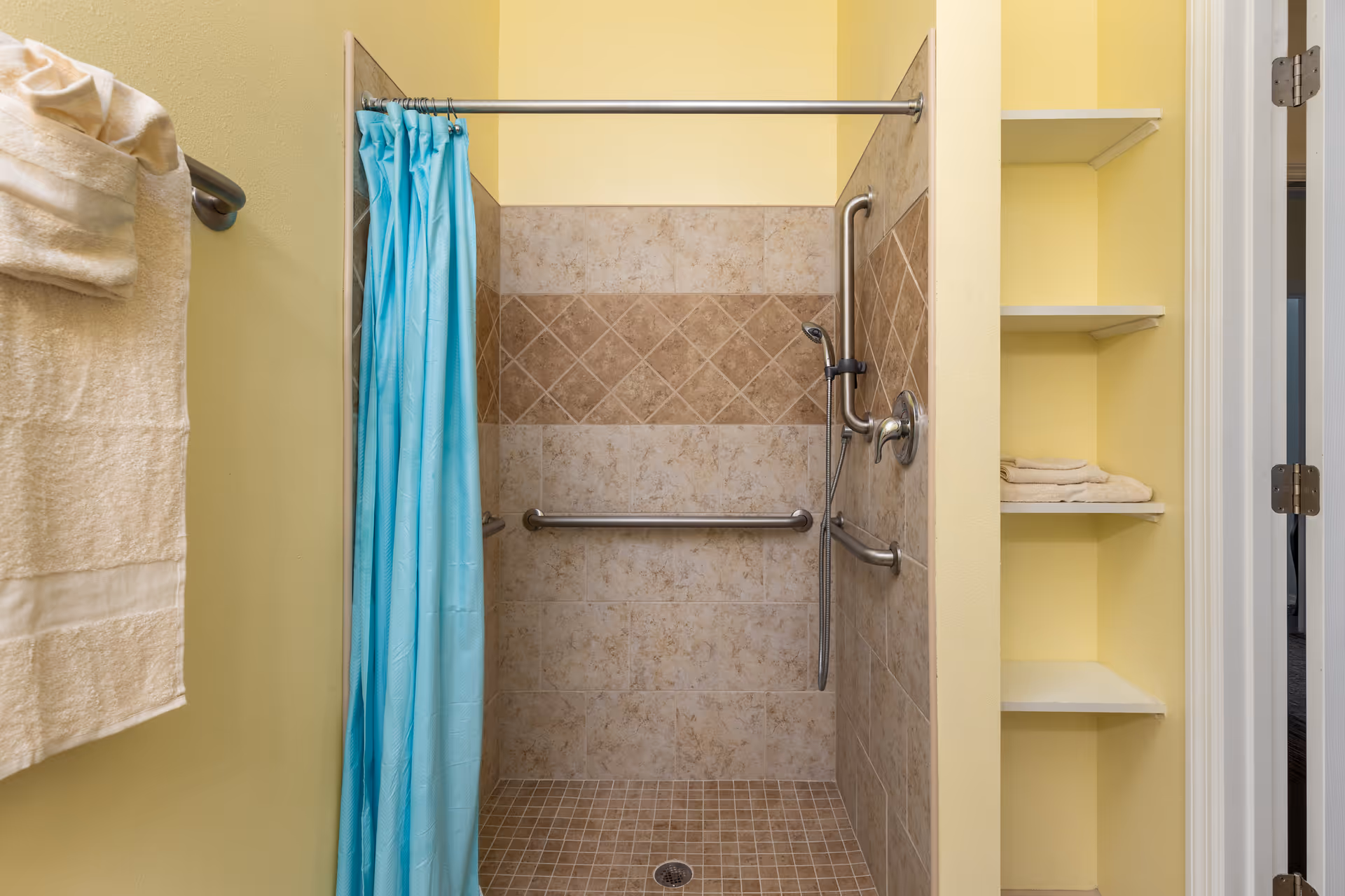 A tiled walk-in shower with beige and brown tiles, equipped with a handheld showerhead and multiple grab bars. A blue shower curtain hangs on a metal rod. To the right, there are built-in shelves with folded towels. On the left wall, a towel rack holds folded beige towels. The walls are painted yellow.