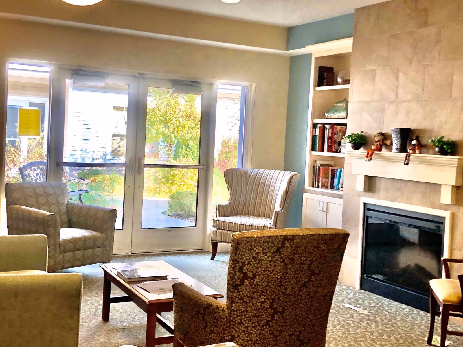 A cozy living room area with several upholstered armchairs arranged around a wooden coffee table. There is a fireplace with a mantel decorated with small plants and figurines. Built-in shelves with books and decorative items are next to the fireplace. Large glass doors lead outside to a garden area with greenery visible through the glass.