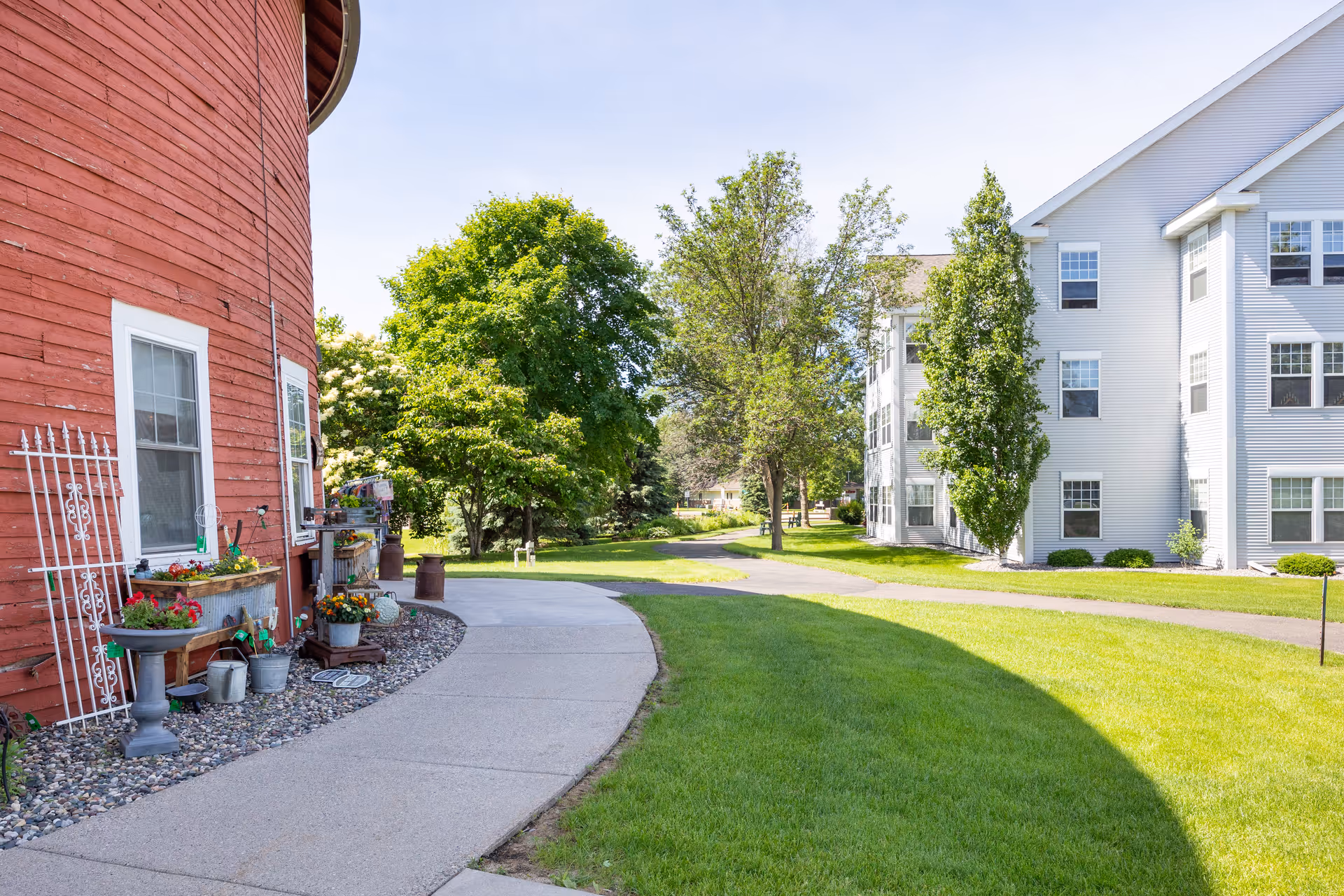 Curved concrete pathway beside a large red barn-like building with flower pots and garden decorations, leading towards a white multi-story residential building surrounded by green grass and trees under a clear sky.