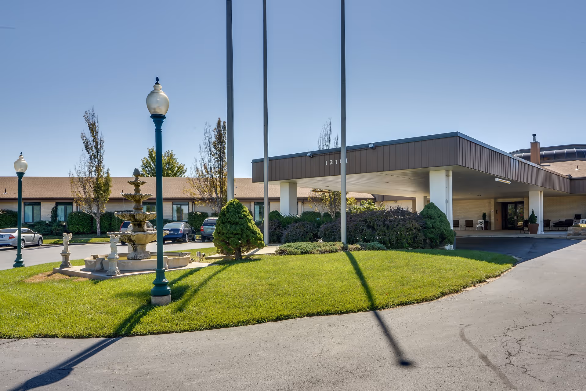 Exterior view of SummitView Terrace senior living facility showing the entrance with a covered drop-off area, a green lawn with a multi-tiered fountain, lamp posts, and several parked cars under a clear blue sky.