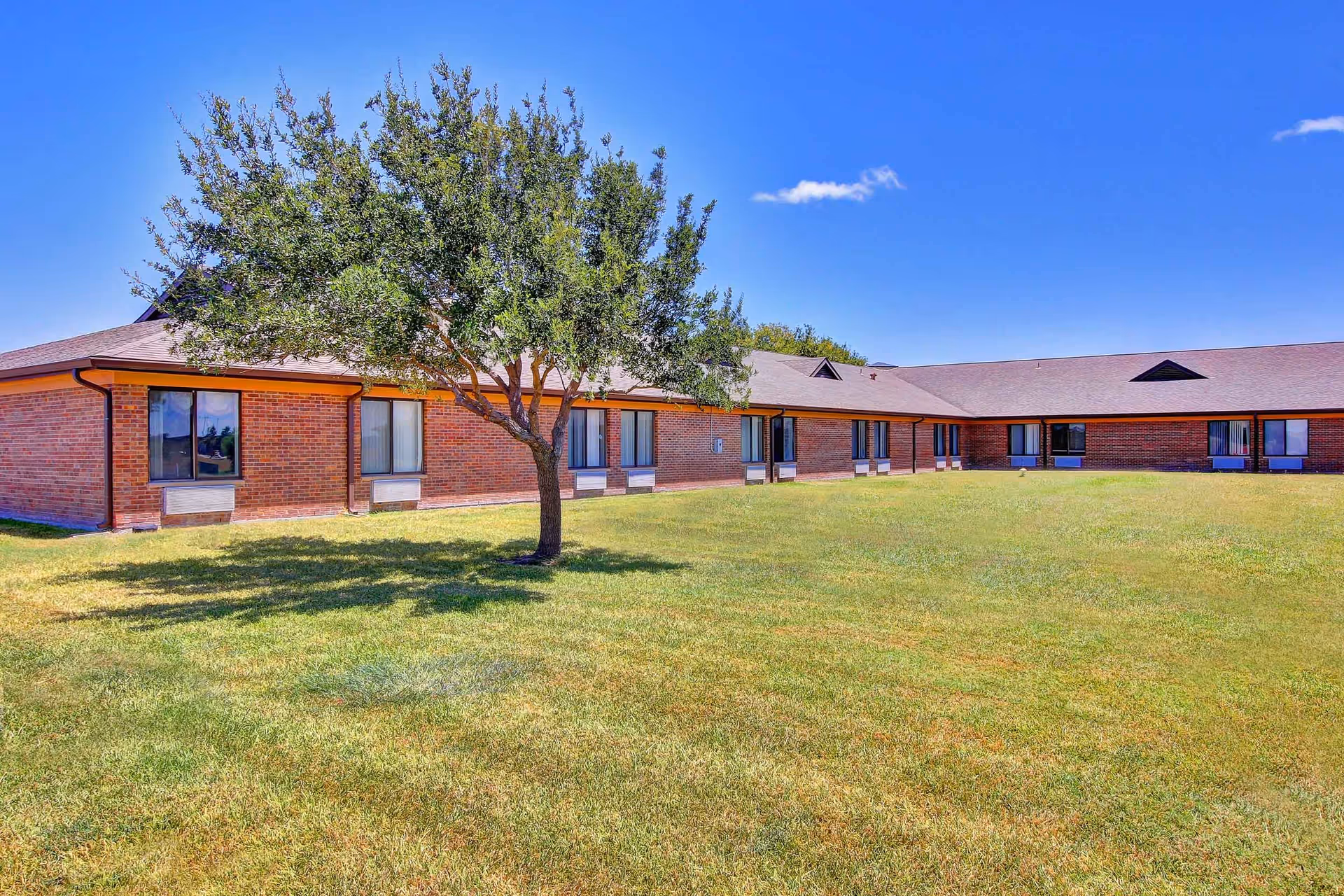 A single-story brick building with multiple windows, surrounded by a large grassy lawn and a tree in the foreground under a clear blue sky.