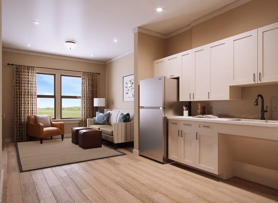 A bright and cozy living area combined with a kitchen. The living area features a window with patterned curtains, a brown armchair, a beige sofa with blue and white pillows, a floor lamp, and two brown ottomans on a beige rug. The kitchen area has white cabinets, a stainless steel refrigerator, a sink with a black faucet, and a countertop with kitchen utensils and a book.