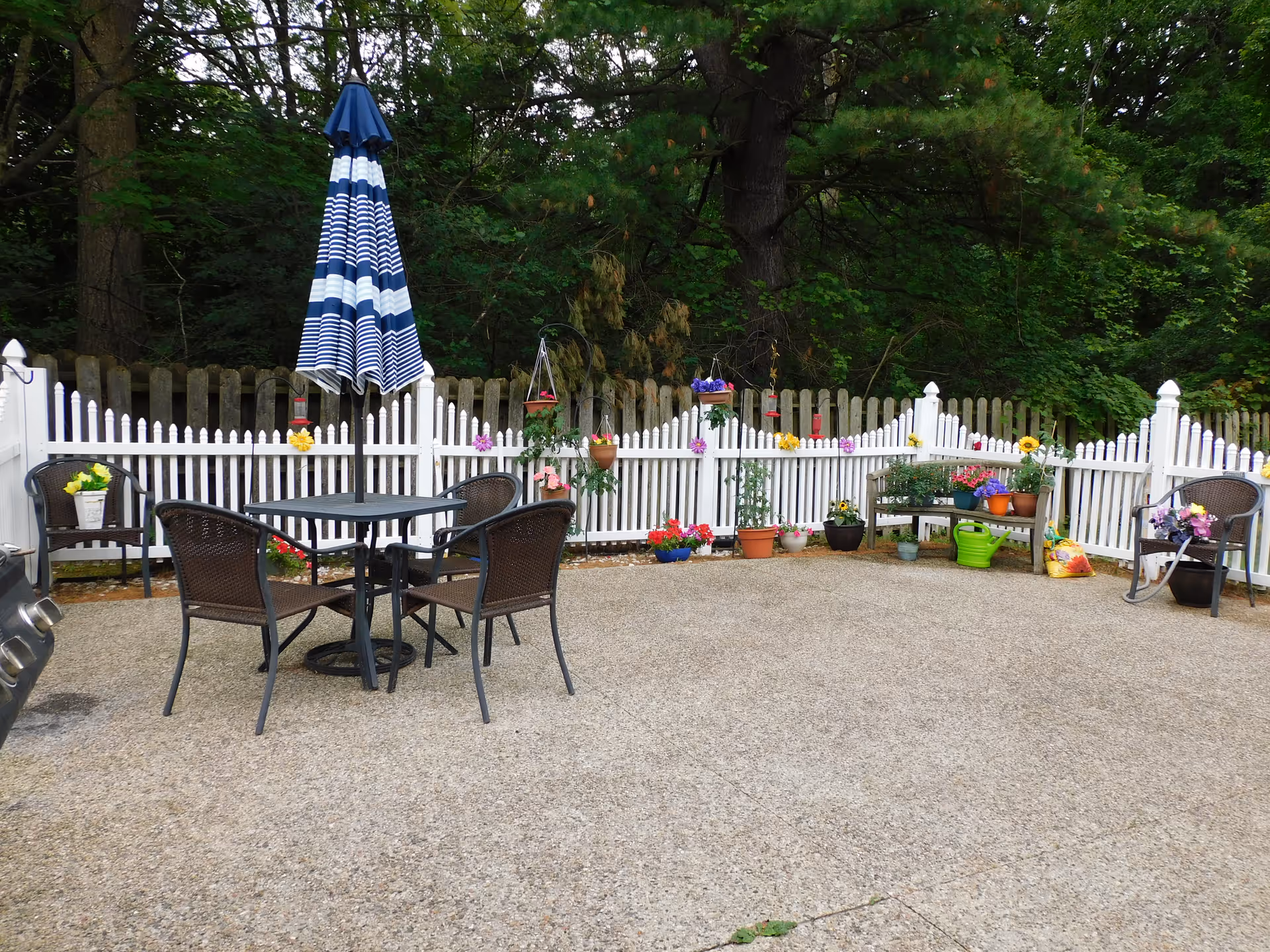 Outdoor patio area with a round table and four chairs, a closed blue and white striped umbrella, and a white picket fence decorated with colorful flowers and potted plants. There is a small bench with more potted plants and gardening supplies against the fence, surrounded by green trees in the background.
