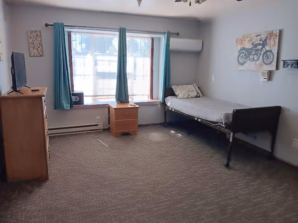 A simple bedroom with a single bed covered with a gray blanket and a pillow, a wooden dresser with a TV on top, a small wooden nightstand under a large window with teal curtains, a wall-mounted air conditioning unit, and a motorcycle-themed wall art.