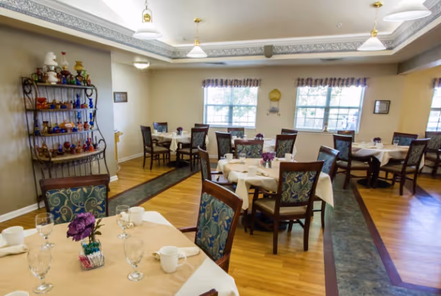 Dining room with multiple tables set with tableware, patterned chairs, wood flooring, decorative shelving, and windows.