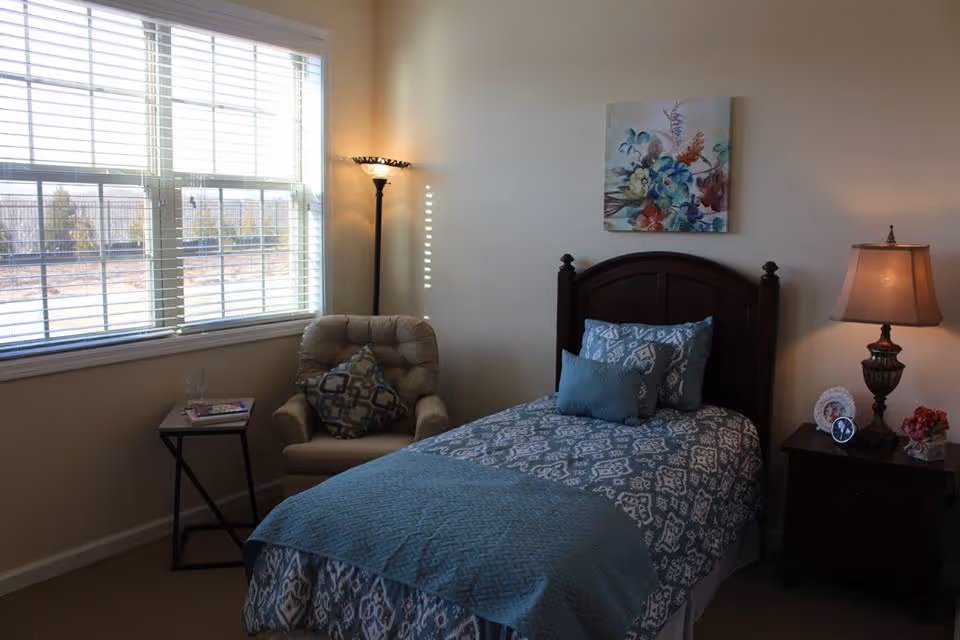 A cozy bedroom with a single bed covered in a blue and white patterned bedspread and pillows. Next to the bed is a wooden nightstand with a lamp, a clock, a small photo frame, and a flower arrangement. A cushioned armchair with a patterned pillow sits near a window with white blinds, and a small side table holds a glass and a book. A floral painting hangs on the wall above the bed.