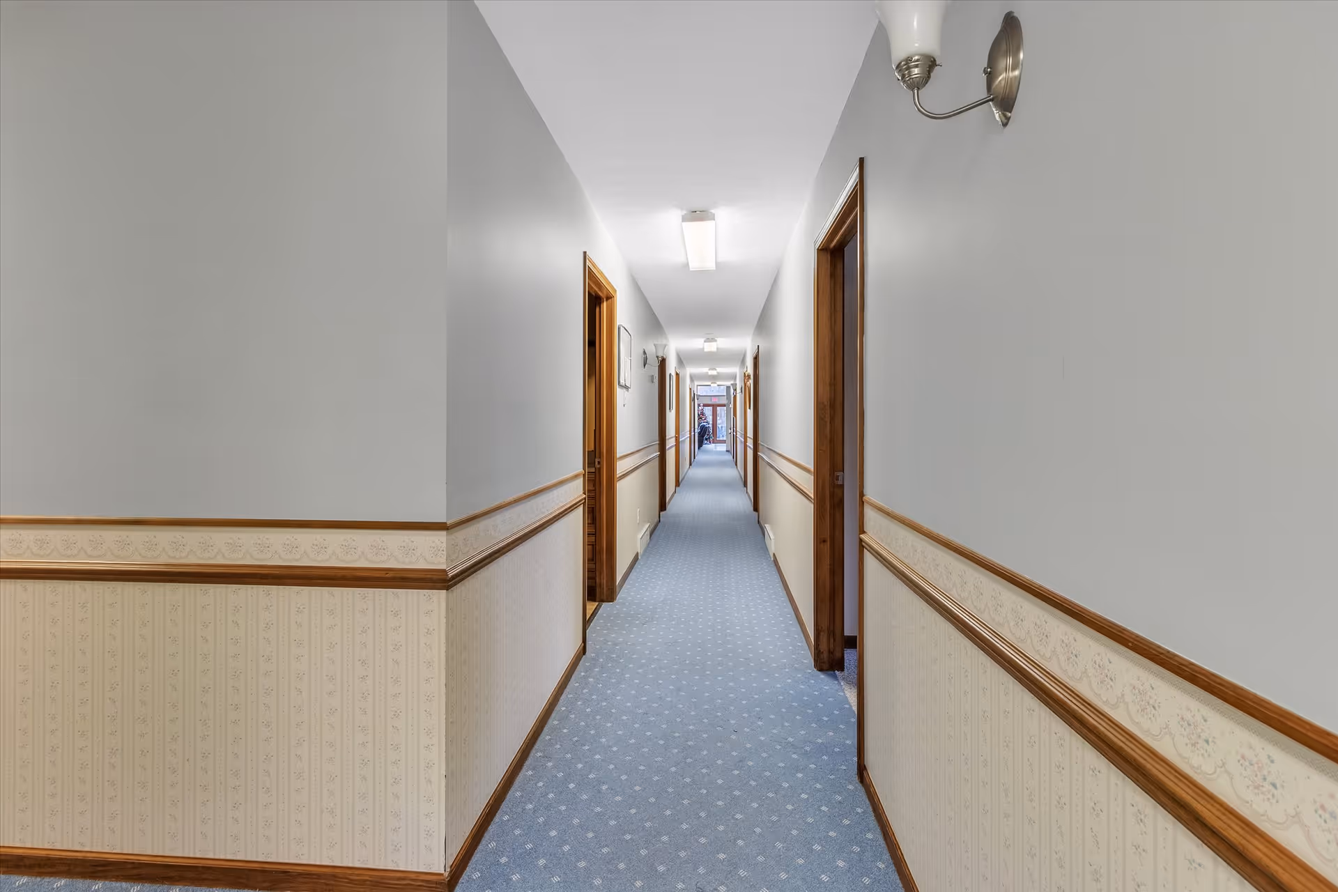 A long, narrow hallway in an adult care home with light gray walls, wooden trim, and blue carpet with a subtle pattern. Several doorways line both sides of the hallway, and ceiling lights illuminate the space.