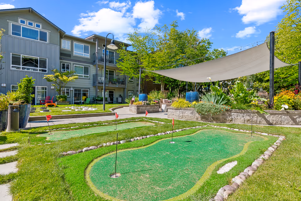 Outdoor area of a senior living facility featuring a small putting green with several holes marked by red flags. Surrounding the green are well-maintained grassy areas, a paved walkway, and landscaped garden beds with various plants and flowers. In the background, there is a three-story residential building with balconies and large windows. A large shade sail is stretched over part of the garden area, and a streetlamp is visible near the building. The sky is clear with some scattered clouds.