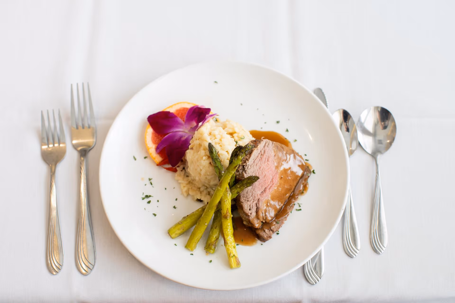 Plated steak with asparagus, rice and a purple flower garnish on a white plate set on a white tablecloth with forks and spoons.