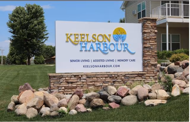 White entrance sign reading 'Keelson Harbour' mounted on a stone base with landscaping and the senior living building behind it.