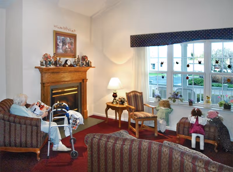 A cozy living room in a senior living facility with an elderly woman sitting on a striped armchair reading a magazine next to a walker. The room features a wooden fireplace mantel decorated with figurines and a framed picture above it. There is a side table with a lamp and a striped chair near a large window with several dolls placed on the window seat. The carpet is red, and the walls are light-colored.