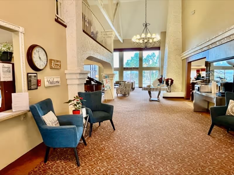 A spacious and well-lit common area in a senior living facility with high ceilings and large windows letting in natural light. The room features blue armchairs with decorative pillows, a round table with flowers, a chandelier, and a piano. There are welcome signs and a clock on the wall, and the carpet has a patterned design.