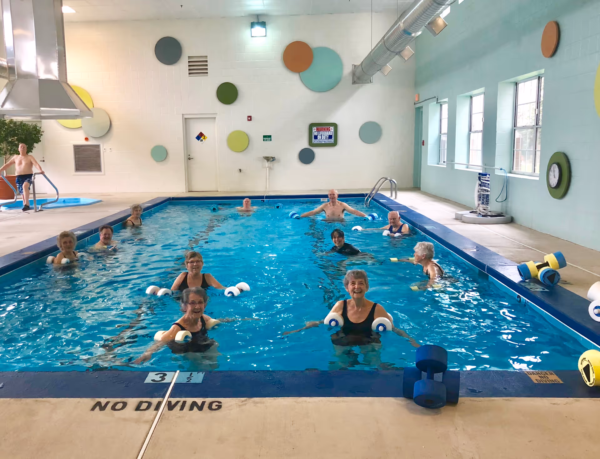 A group of elderly people participating in a water exercise class in an indoor swimming pool. The pool area has colorful circular wall decorations, large windows on one side, and various pool exercise equipment like foam dumbbells and noodles. The floor near the pool edge has a 'NO DIVING' warning.