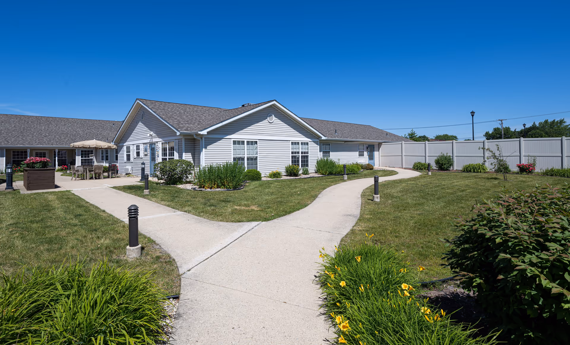 Outdoor view of a single-story white building with a gray roof, surrounded by a well-maintained lawn and garden beds with green shrubs and yellow flowers. A concrete walkway splits into two paths leading to the building entrance and a patio area with tables, chairs, and an umbrella. The sky is clear and blue.