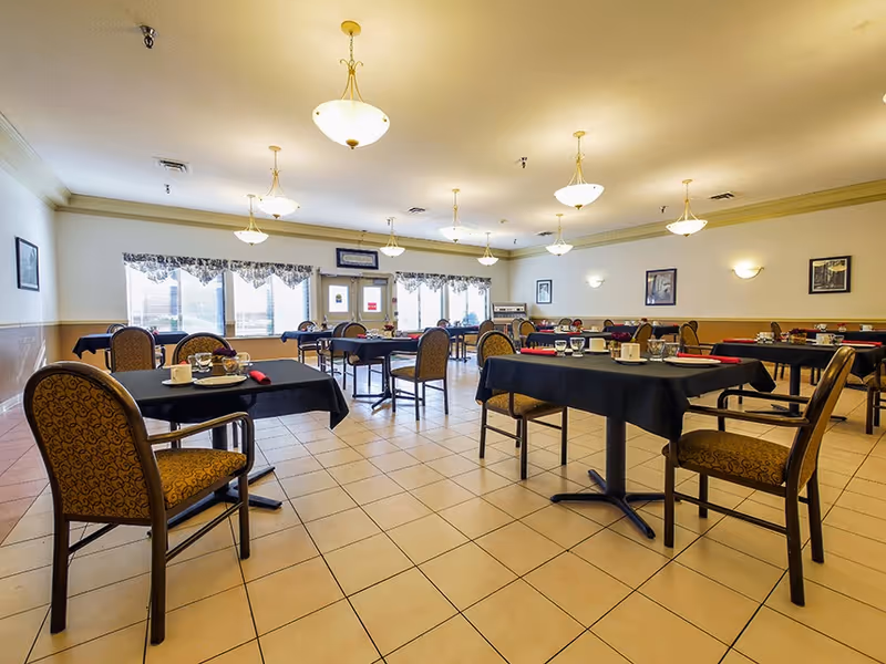 A spacious dining room with multiple tables covered in black tablecloths, each set with cups, glasses, and napkins. The room has tiled floors, patterned chairs, large windows with valances, and ceiling lights providing warm illumination.