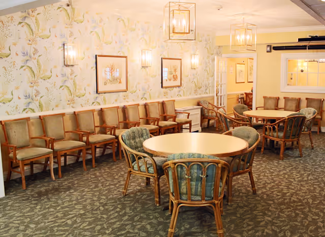 A well-lit common area with round tables surrounded by cushioned wicker chairs and a row of wooden chairs with armrests lined up against a wall with floral wallpaper and framed artwork. The room has carpeted flooring and hanging lantern-style light fixtures.