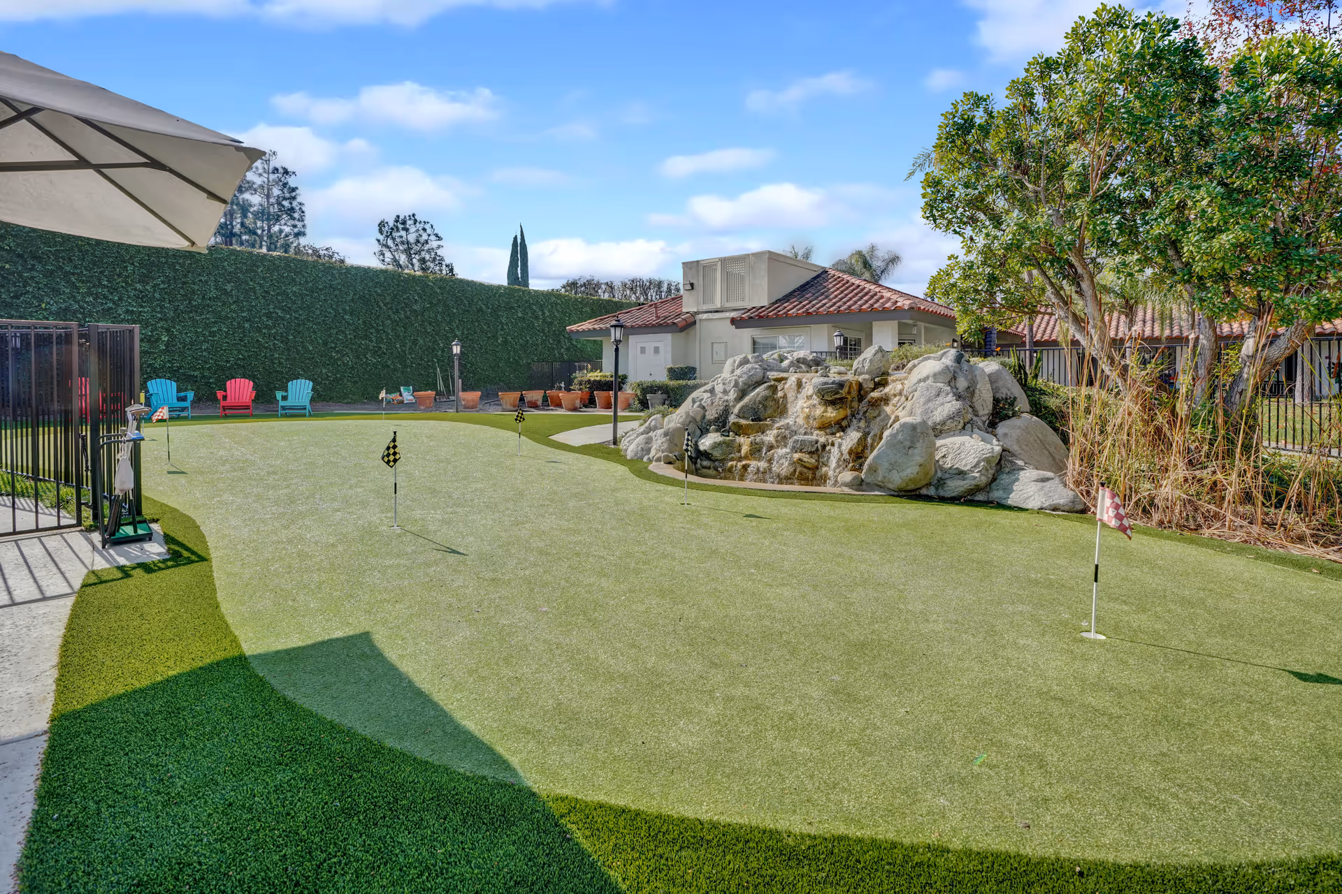 Outdoor putting green with small flags, a rock waterfall feature, colorful chairs and a tiled-roof building under a blue sky.