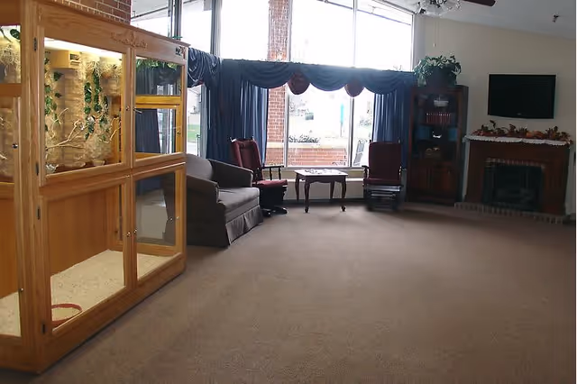 Spacious common room with armchairs, a couch and table by large windows, a fireplace and TV, and a wooden terrarium on the left.