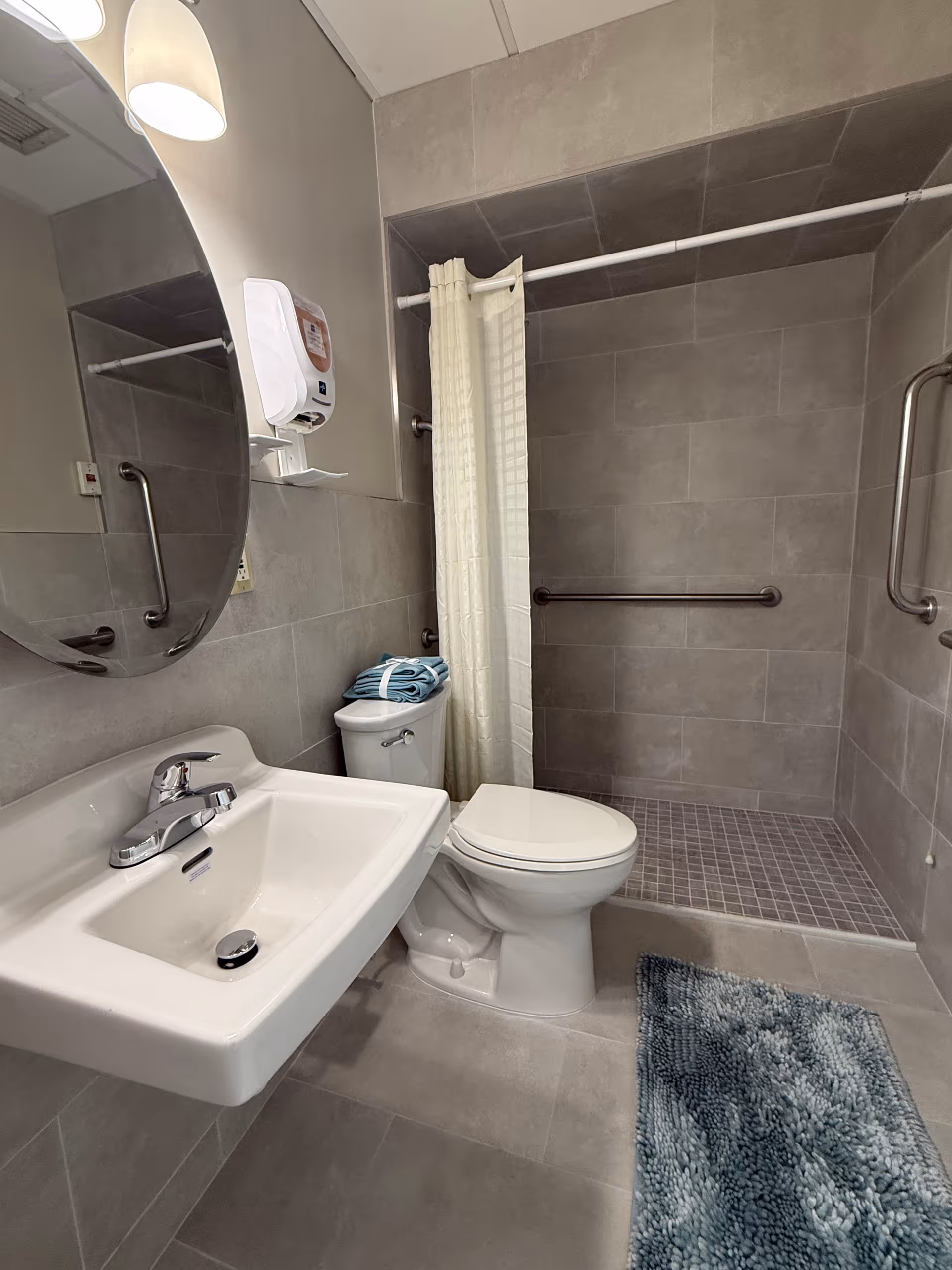 A modern bathroom with gray tiled walls and floor, featuring a white wall-mounted sink with a chrome faucet, a round mirror above the sink, a white toilet with folded blue towels on top, and a walk-in shower with a white curtain and grab bars on the walls. A blue textured bath mat is placed on the floor in front of the shower.