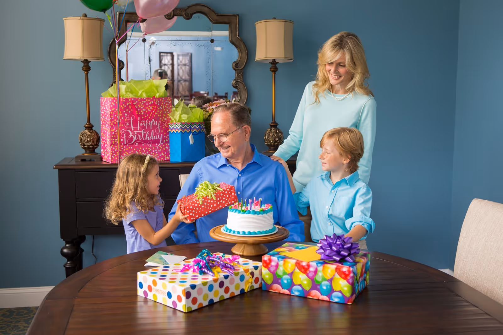 An elderly man sitting at a dining table with a birthday cake in front of him, surrounded by a young girl handing him a gift, a young boy, and a woman standing behind them, all smiling. The table has several wrapped presents and party decorations. In the background, there is a sideboard with more gifts and balloons, and a large mirror on the wall.