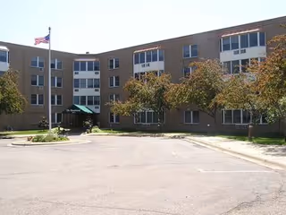 Exterior view of a three-story senior living facility building named Crest View - The Boulevard with an American flag on a flagpole, trees, and a paved driveway in front.