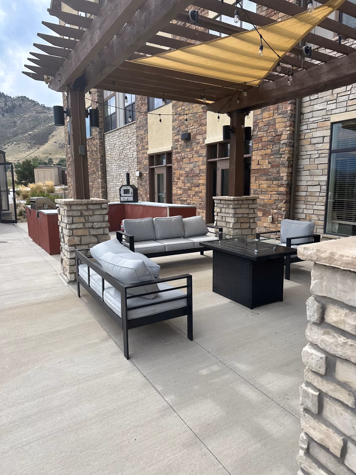 Outdoor patio area with cushioned seating including a sofa and chairs arranged around a black fire pit table. The patio is covered by a wooden pergola with string lights and a fabric shade. The building exterior features stone and brick walls with windows and doors. In the background, there are hills and some greenery.