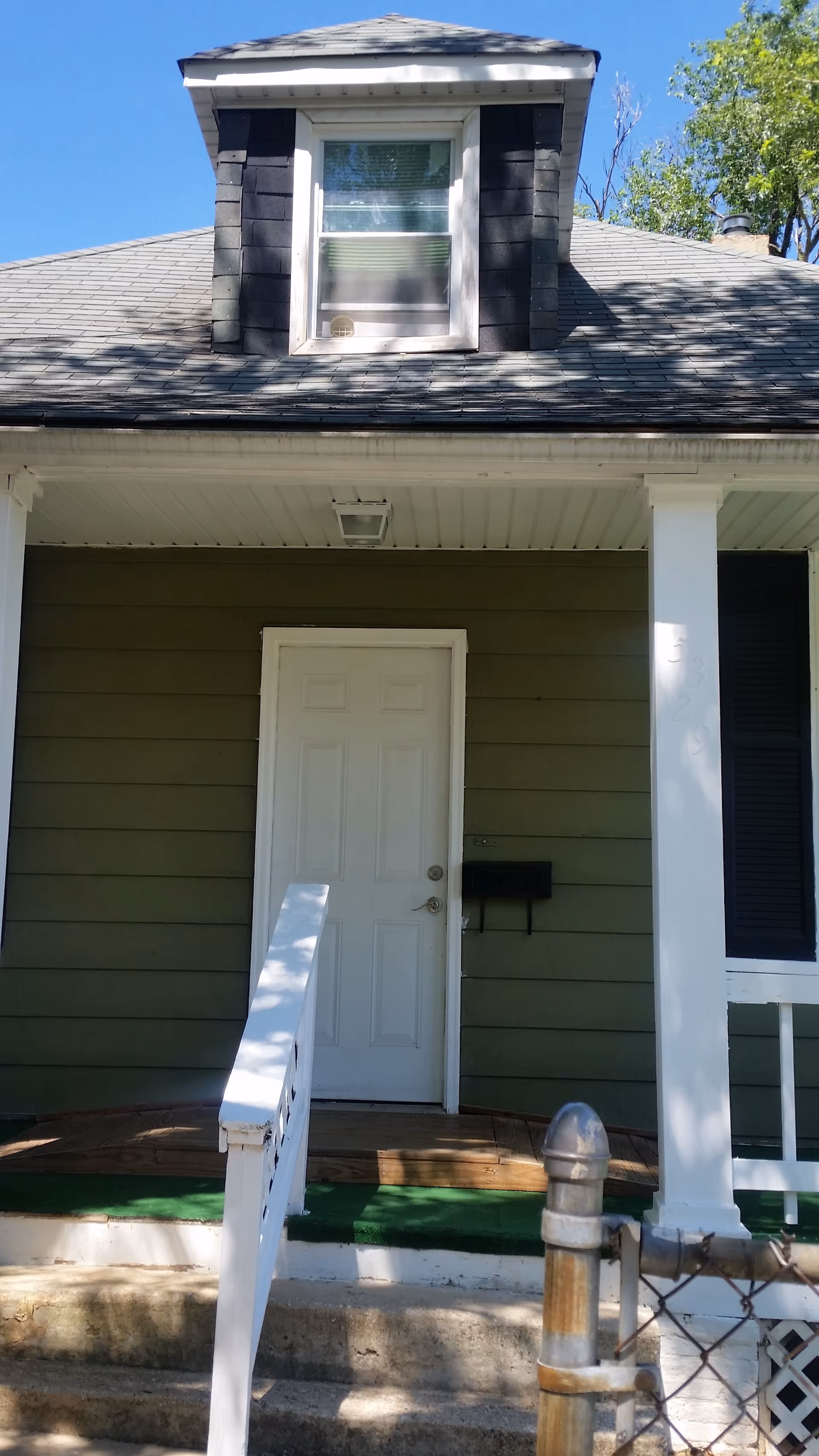 Front entrance of a small house with green siding, a white door and porch with railing, and a dormer window above.