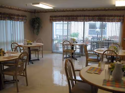 A bright senior living dining room with round tables, chairs, and large windows.