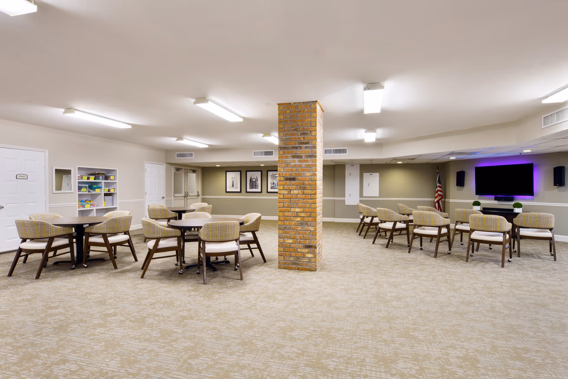 Spacious senior living common room with round tables and chairs, a central brick column, and a wall-mounted TV.
