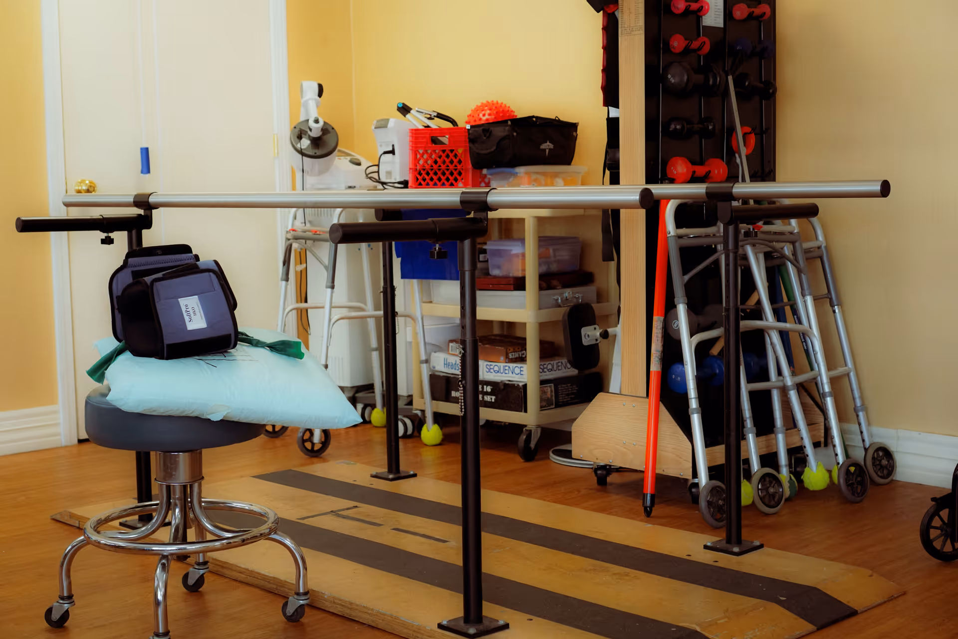 Physical therapy room with parallel bars, walkers, exercise equipment, and a stool with a pillow.