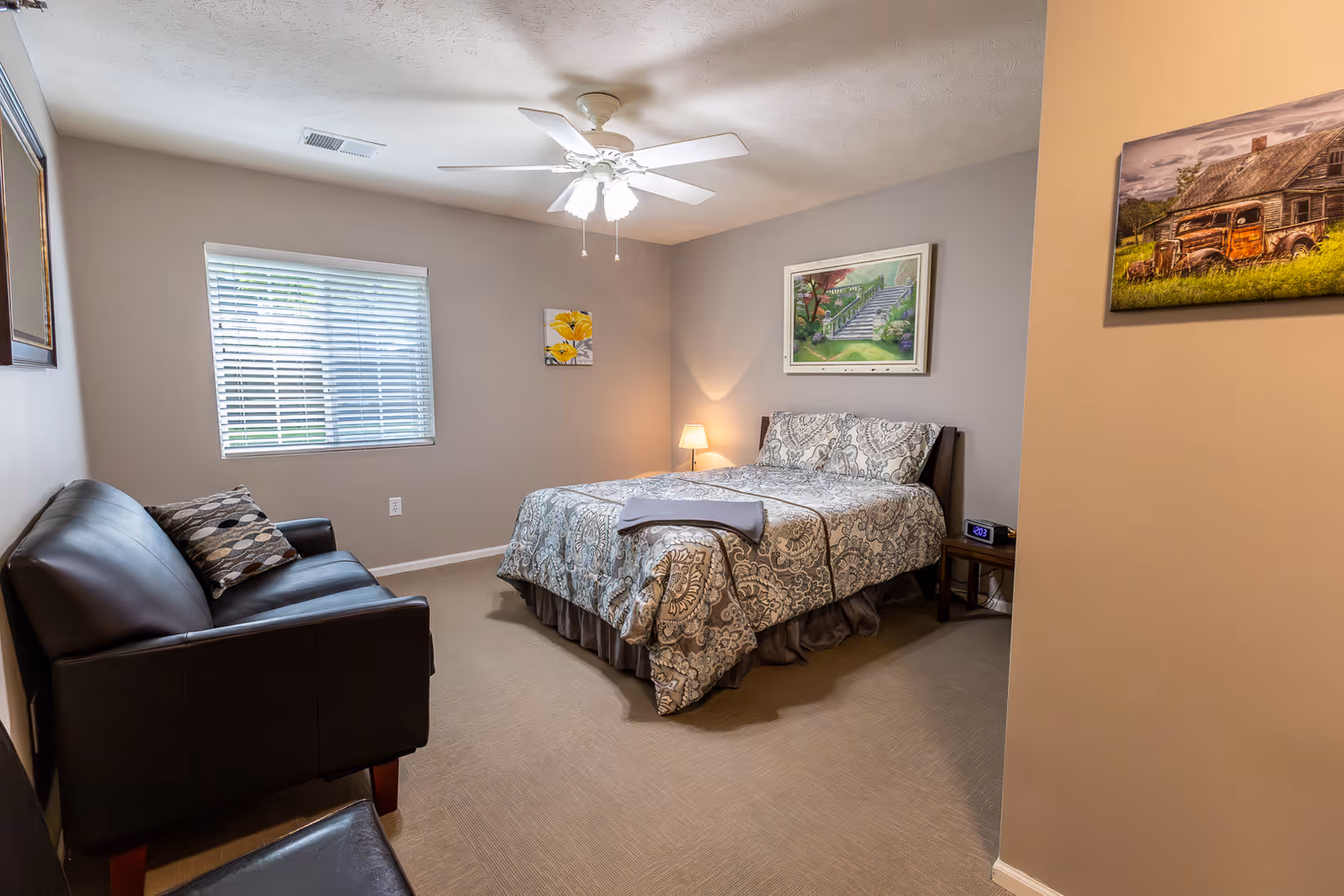 A bedroom with a bed covered in patterned bedding, a small bedside table with a lamp and digital clock, a black leather couch with a decorative pillow, a window with blinds, ceiling fan with lights, and framed artwork on the walls.
