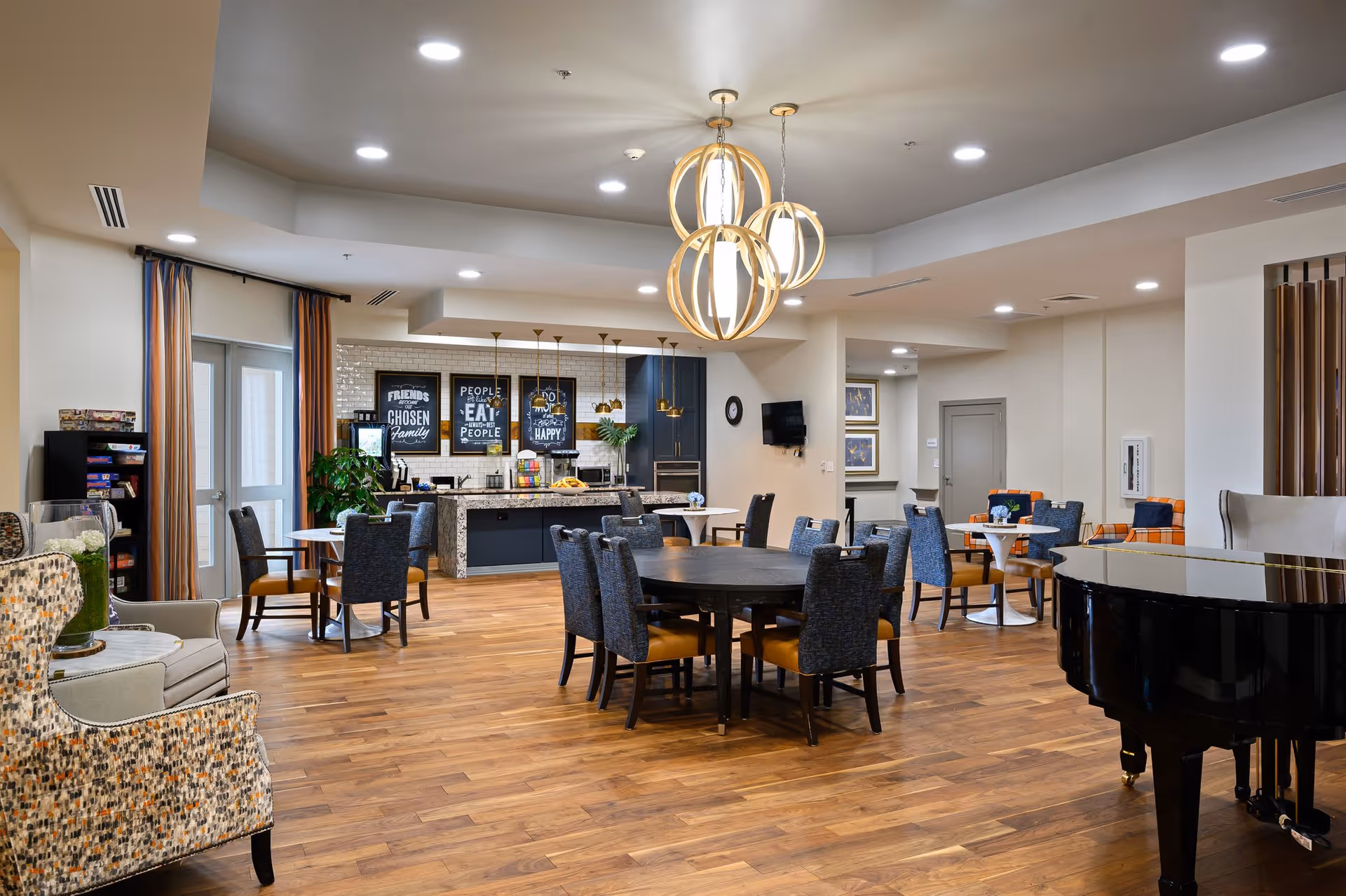 A spacious senior living common area with wooden flooring, multiple round tables surrounded by chairs, a black grand piano, and a kitchen area in the background with a granite countertop and decorative wall art. The room is well-lit with modern ceiling lights and pendant lamps, and there are comfortable armchairs and a small bookshelf on the left side.