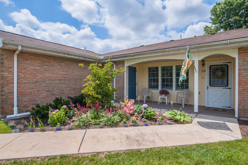Front porch of a senior living community unit with a small garden bed featuring various flowers and plants. The porch has two chairs and a small table with a flower pot. A decorative flag is hanging near the entrance door, which has a wreath. The building exterior is brick and light-colored siding under a partly cloudy sky.