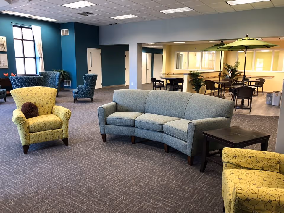 A spacious and well-lit common area with a light blue curved sofa, two yellow patterned armchairs, and several blue armchairs in the background. The room has a carpeted floor, teal and light blue walls, and large windows letting in natural light. In the background, there is a seating area with tables, chairs, and green umbrellas, along with some potted plants.