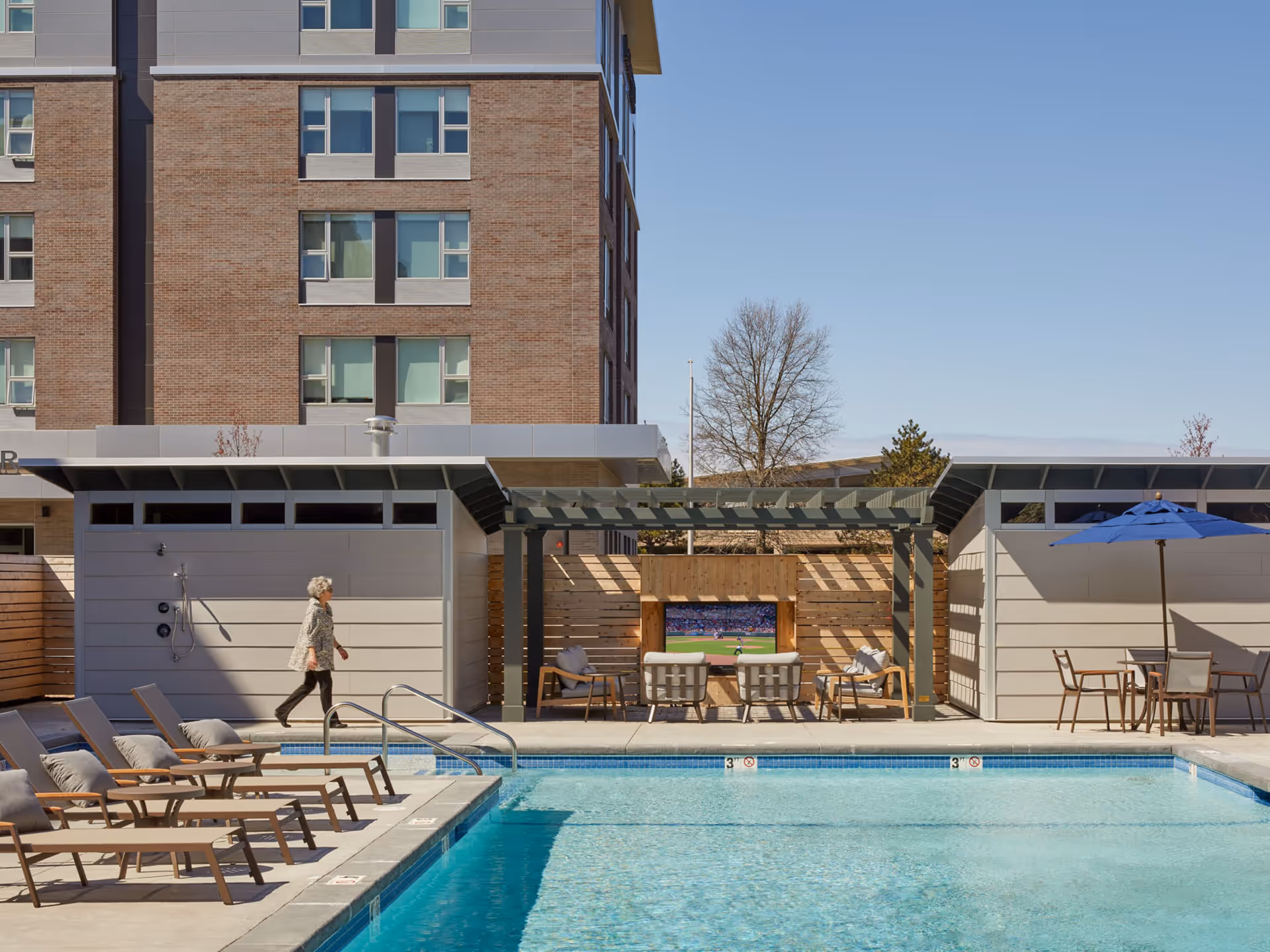 Outdoor pool area at a senior living facility with lounge chairs lined up along the poolside, a pergola with seating and a TV, a woman walking near the pool, and a building in the background under a clear blue sky.