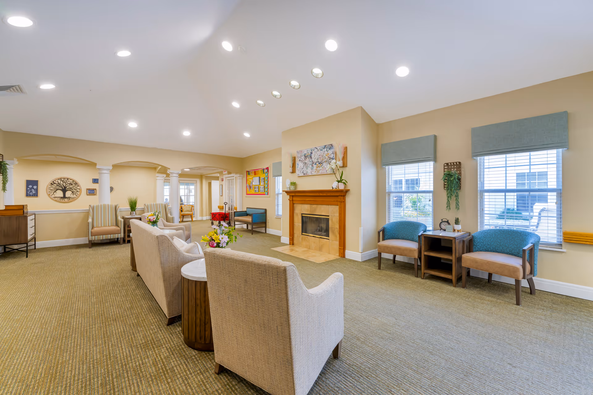 A spacious and well-lit living room area in a senior living facility with beige walls and carpet. The room features a fireplace with a wooden mantel, several upholstered chairs and sofas arranged for conversation, side tables with flowers, large windows with blue valances, and decorative wall art including a tree sculpture and a colorful painting.