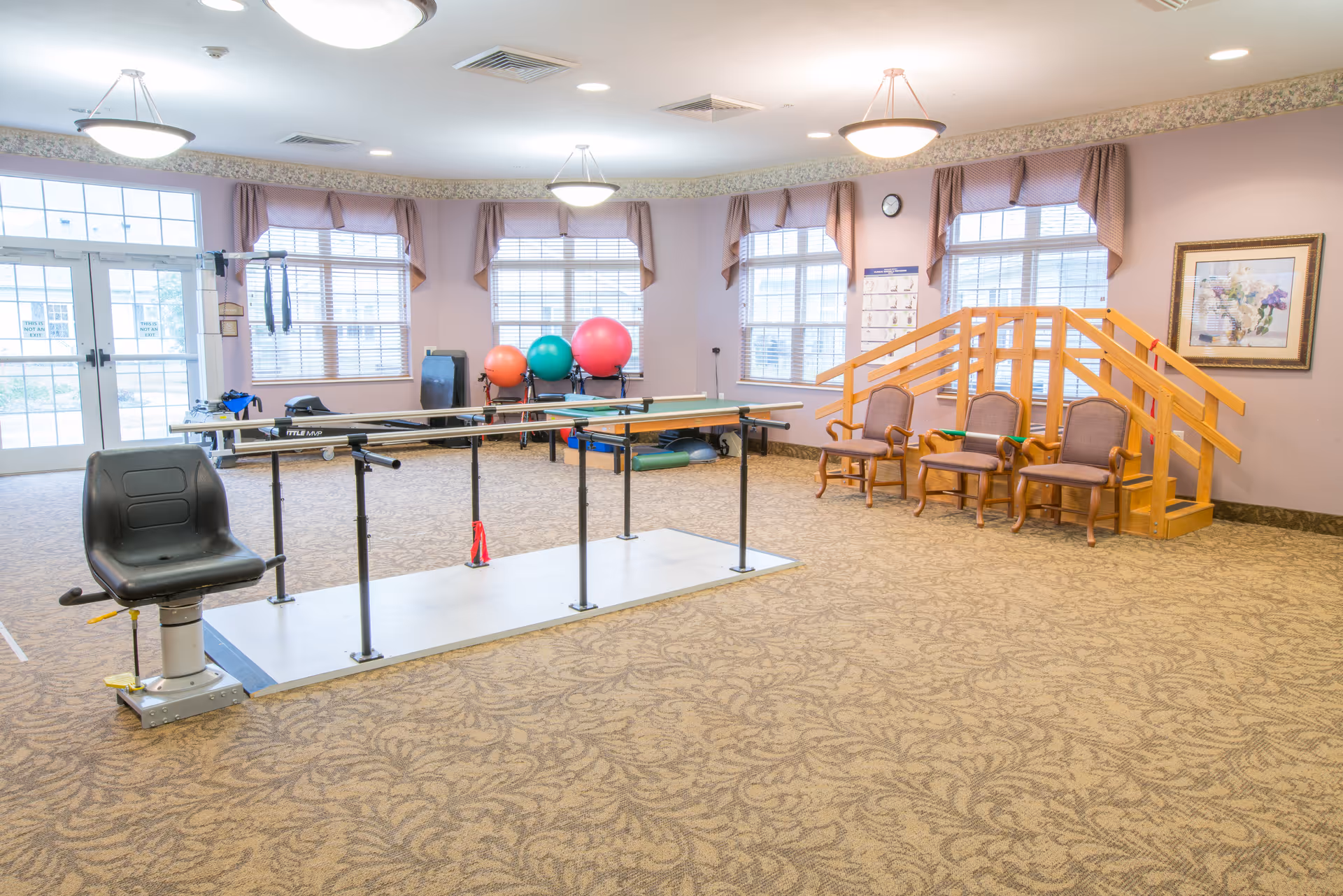A spacious rehabilitation room with parallel bars for walking exercises, a black chair attached to the bars, three wooden chairs with armrests, a small wooden staircase with handrails, and several large exercise balls near windows with blinds. The room has beige patterned carpet, light purple walls, and ceiling lights.