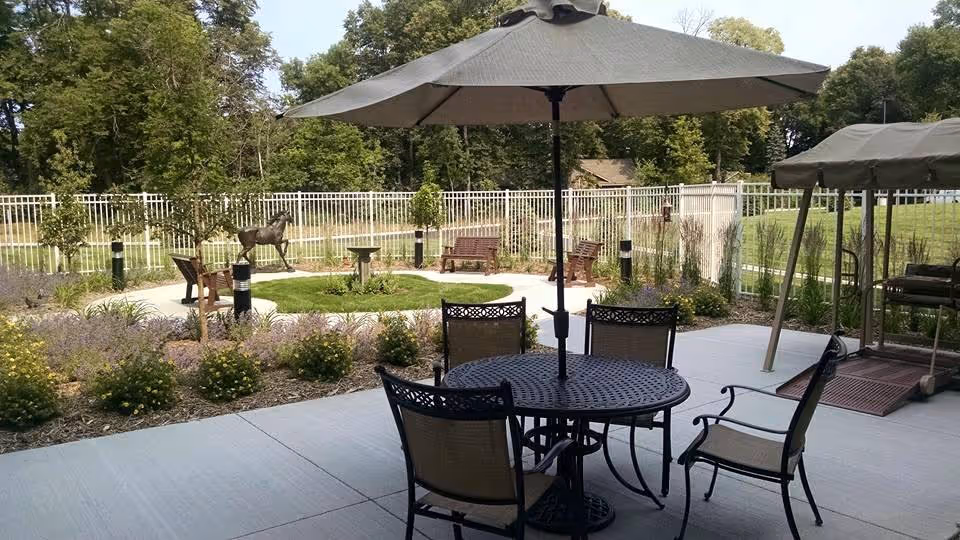 Outdoor patio area with a round metal table and four chairs under a large umbrella. In the background, there is a fenced garden area with benches, a birdbath, and a horse statue surrounded by greenery and trees.
