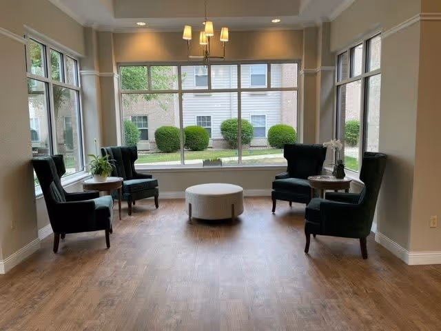 A bright seating area with four dark green upholstered armchairs arranged around two small round wooden tables and a central round ottoman. Large windows provide a view of trimmed bushes and a building exterior, allowing natural light to fill the space. A modern chandelier hangs from the ceiling.