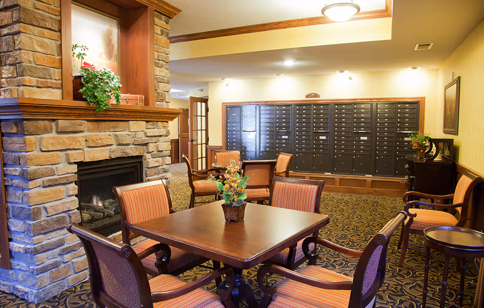 Cozy senior living common room with a stone fireplace, wooden table and chairs and a wall of mailboxes in the background.