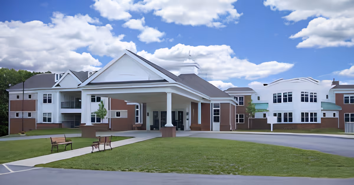 Front exterior view of Plymouth Harborside HealthCare building with a covered entrance, green lawn, benches, and a partly cloudy sky.