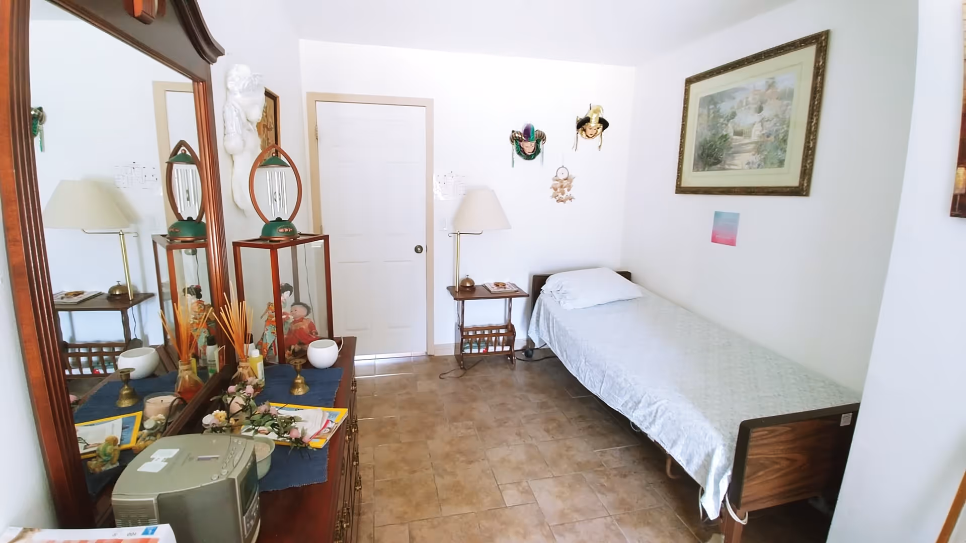 Small bedroom with a single bed along the right wall, a wooden dresser with a large mirror and lamp on the left, and a door at the far end.