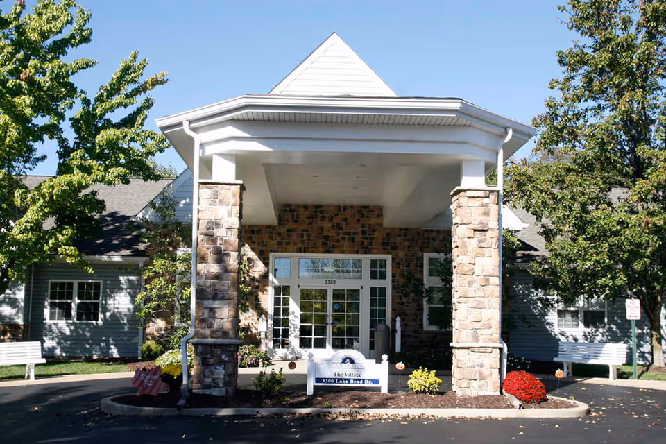 Front exterior view of a senior living facility entrance with stone pillars supporting a covered porch, surrounded by trees and landscaping, with a sign reading 'The Village 3300 Lake Bend Dr.'