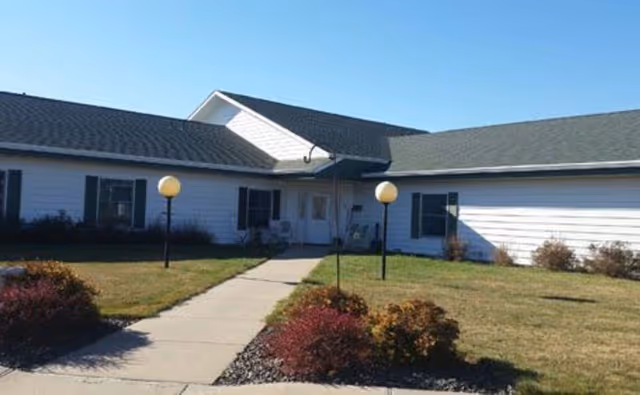 Single-story white assisted living building with a central entrance, walkway, lamp posts, and landscaped lawn under a clear blue sky.